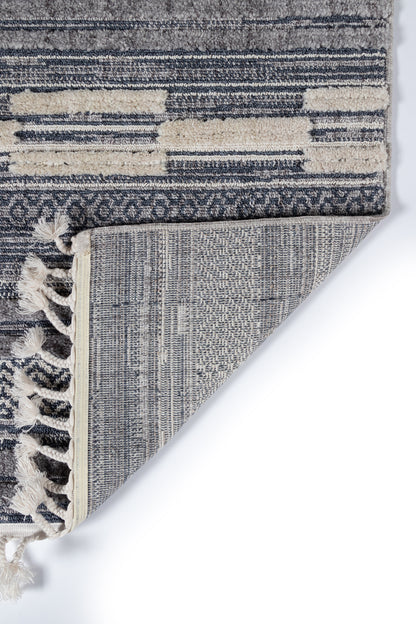 Blue-gray and cream woven area rug with abstract stripe pattern and fringe tassels at edge; folded corner shows underside.