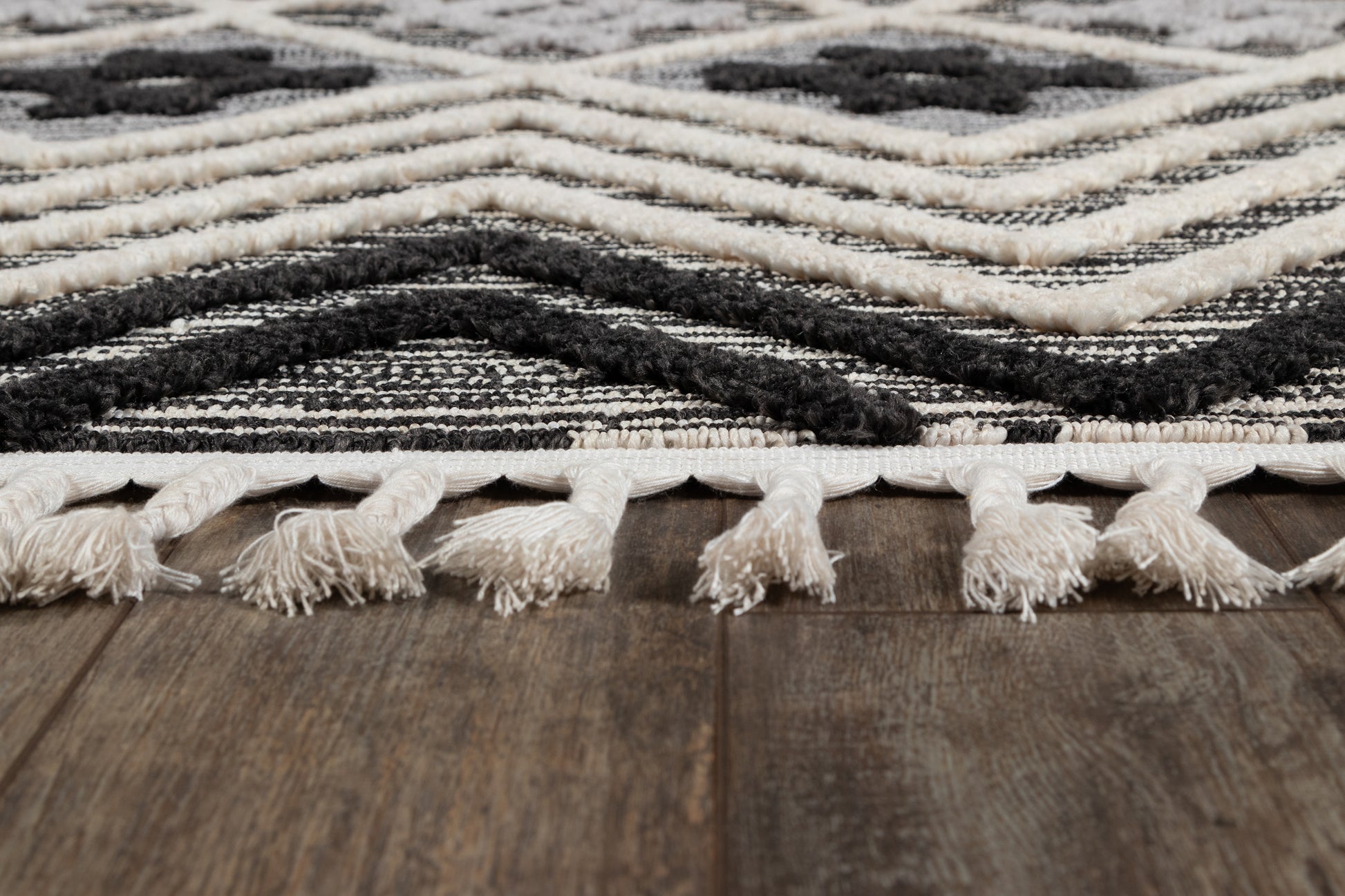 Black and ivory geometric woven rug with white fringe tassels on dark wood floor.