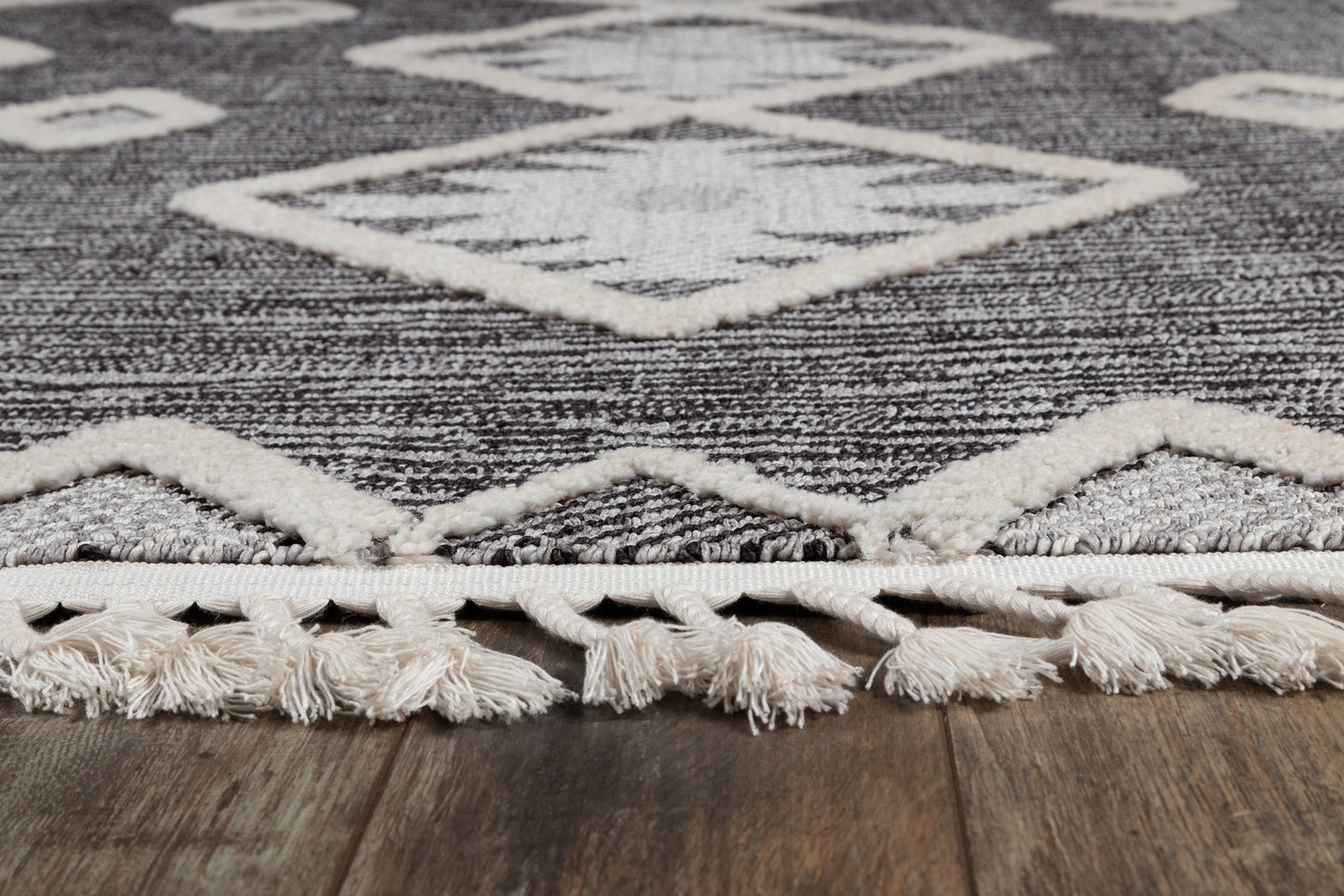 Charcoal gray woven area rug with ivory geometric diamonds and cream fringed edge on wood floor.