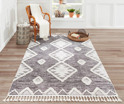 Geometric area rug with white diamond motifs and fringe, charcoal brown tones on a wood floor beside a wicker chair.
