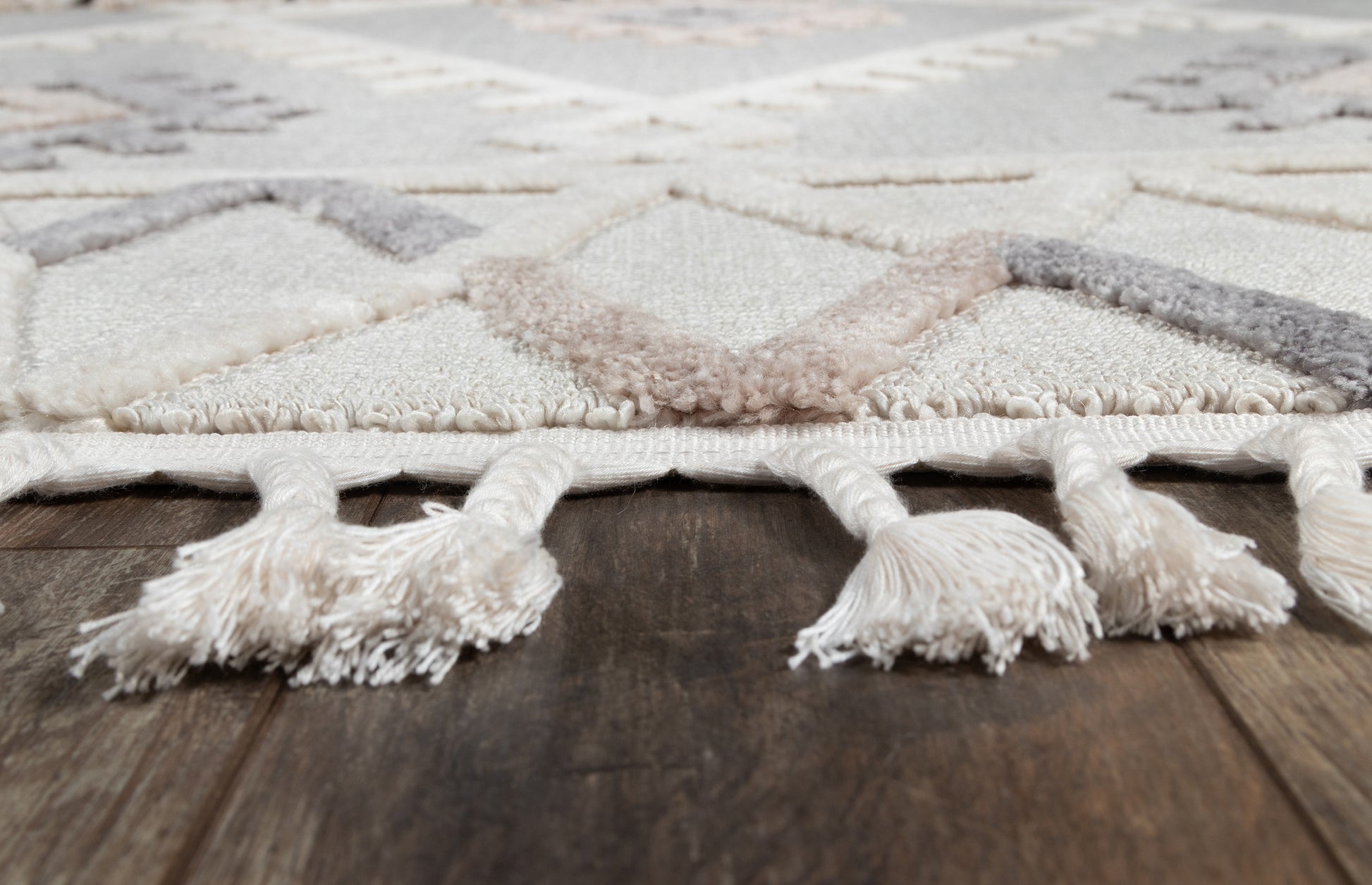Ivory and pale gray geometric rug with diamond motif and fringed tassels along edge on dark wood floor.