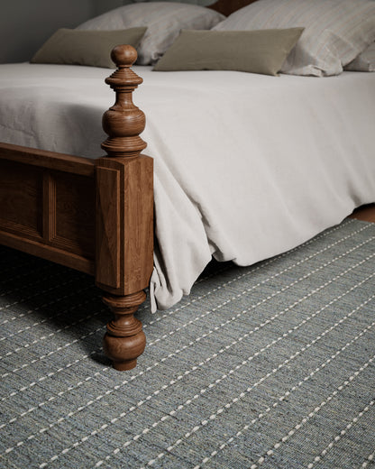 Brown stained wooden bedpost with turned finial; light gray duvet, beige pillows, and a gray striped rug.