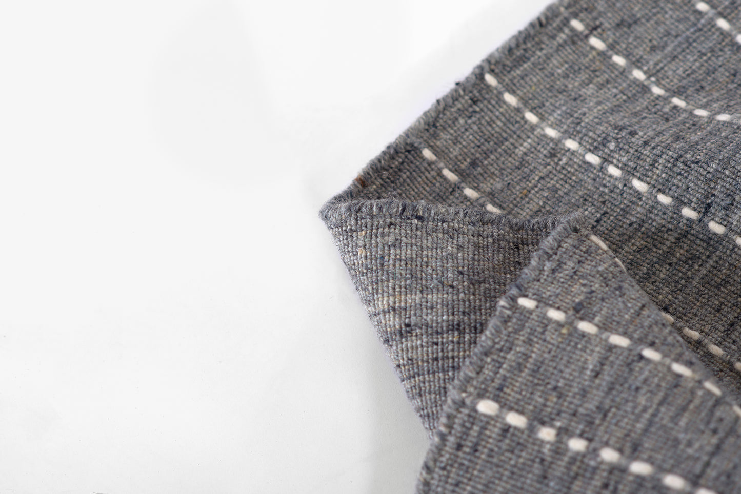 Grey-blue woven rug with white dashed stitch lines, folded corner, textured cotton/linen weave.
