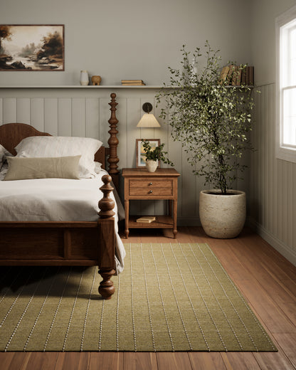 Olive-green striped rug beside a carved wooden bed, nightstand with lamp, and potted tree in a cozy neutral bedroom.