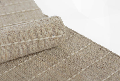 Speckled natural linen blend rug close-up, beige woven texture with white stitched lines and rolled edge.