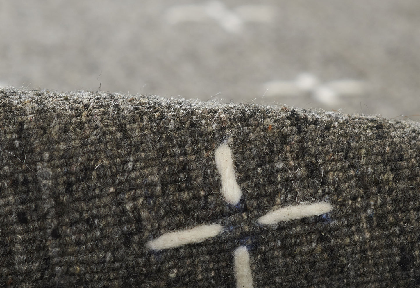 Close-up of olive-gray tweed fabric with white cross-stitch motif; textured wool blend.