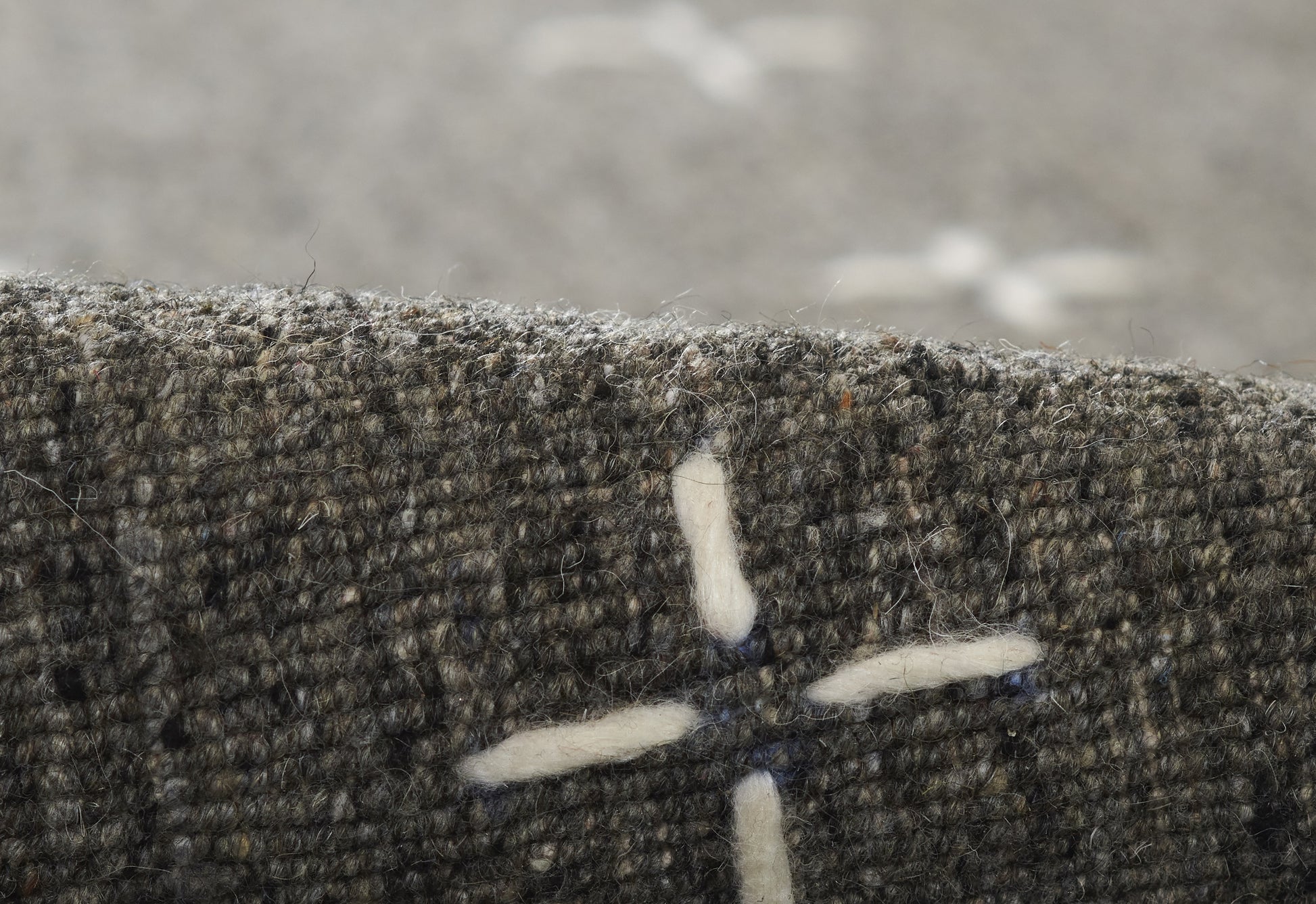 Close-up of olive-gray tweed fabric with white cross-stitch motif; textured wool blend.