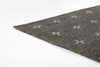 Gray textured rug with white cross-stitch X pattern, close-up corner view.