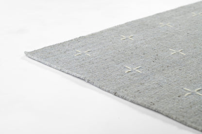 Light gray textured area rug with cream cross-stitch X motifs and fringed edge.