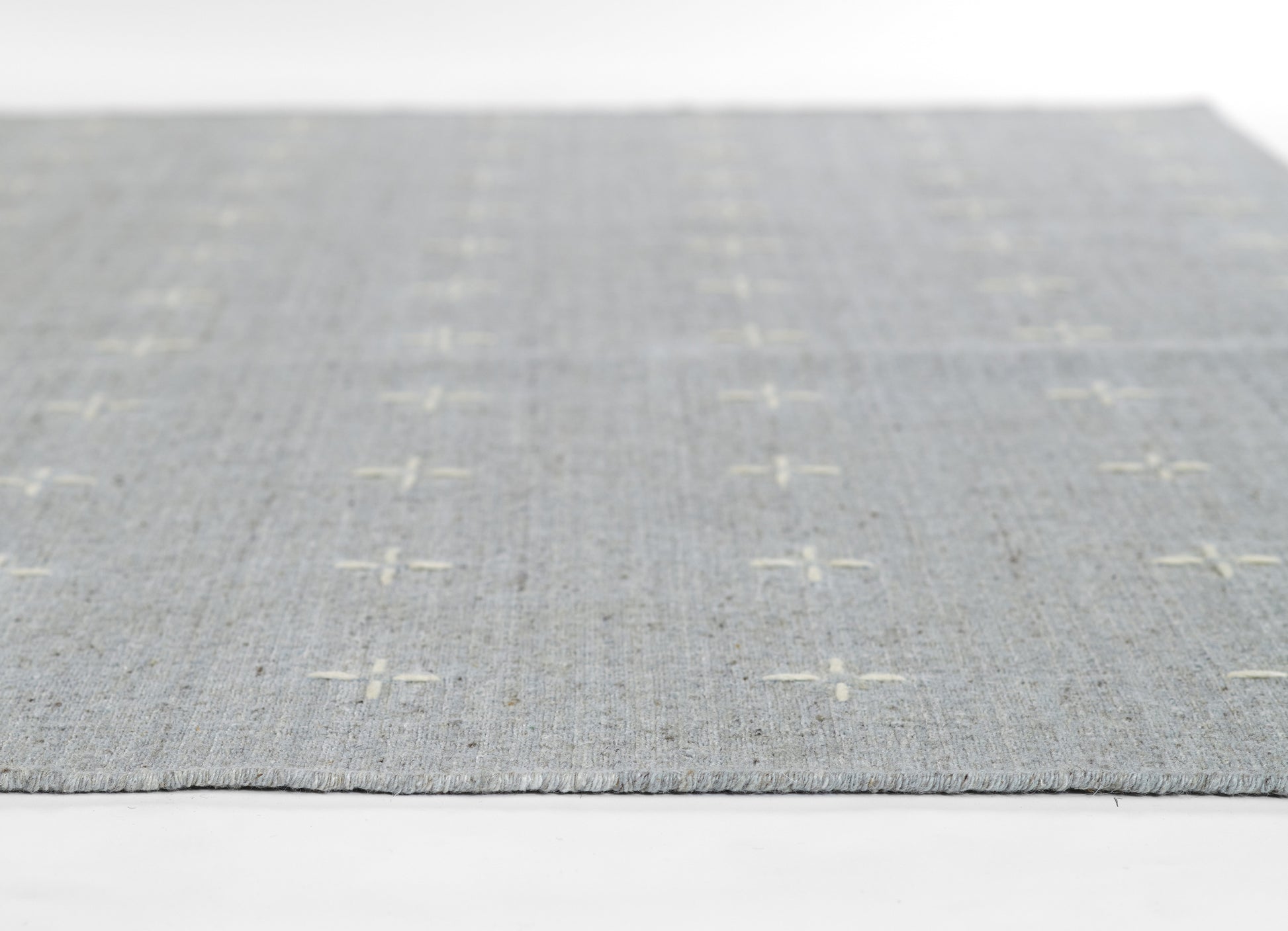 Light gray woven rug with subtle white cross-stitch plus pattern and fringed edges.