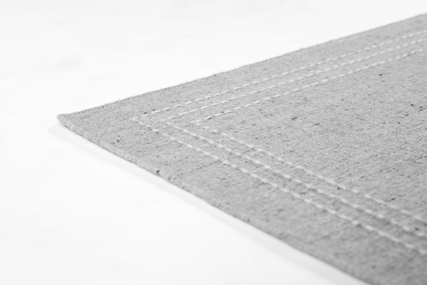 Light gray textured rug corner with double stitched border along the edge.