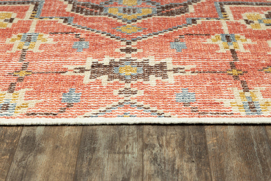 Orange-red geometric tribal rug with blue, yellow and cream motifs on dark wood floor, fringe along bottom edge.