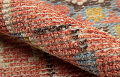 Close-up of a red-toned woven rug with cream, blue and yellow accents; textured looped pile and rolled edge.