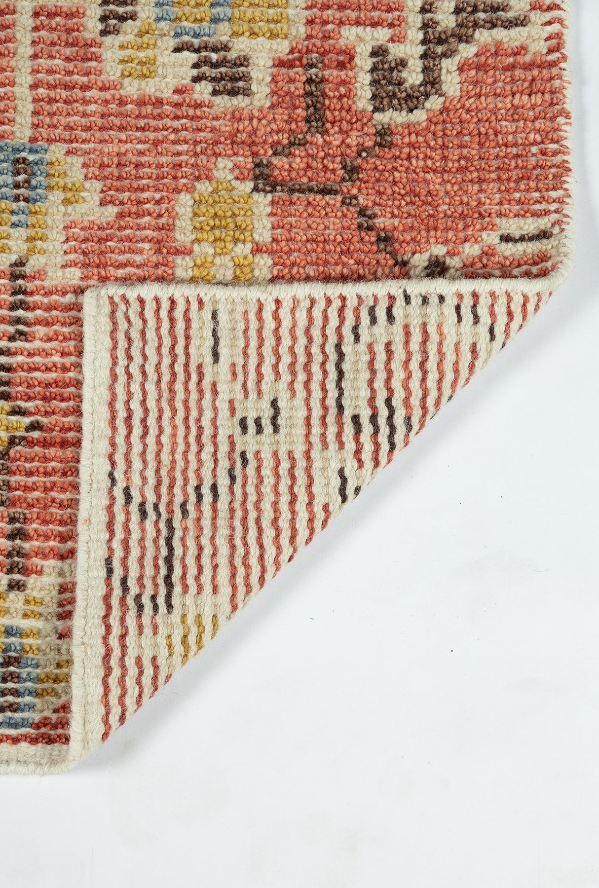 Red-pink woven wool rug with multicolor floral motif; folded corner reveals striped underside.