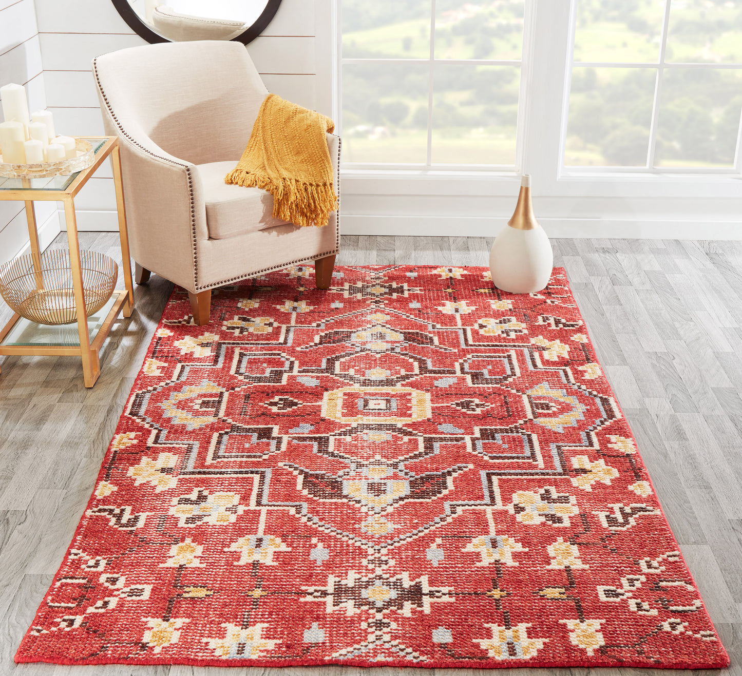 Red geometric area rug with ivory accents in a sunlit living room, beige armchair with orange throw and gold side table.