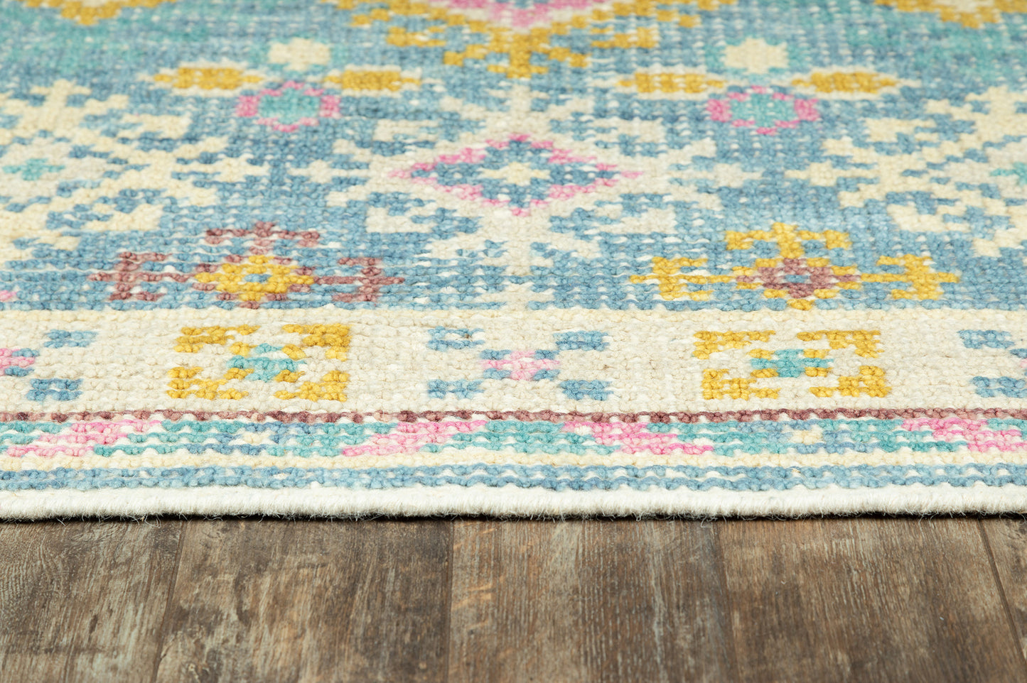 Pastel geometric kilim-style rug with turquoise & pink & yellow motifs on white, fringed edge on wood floor.