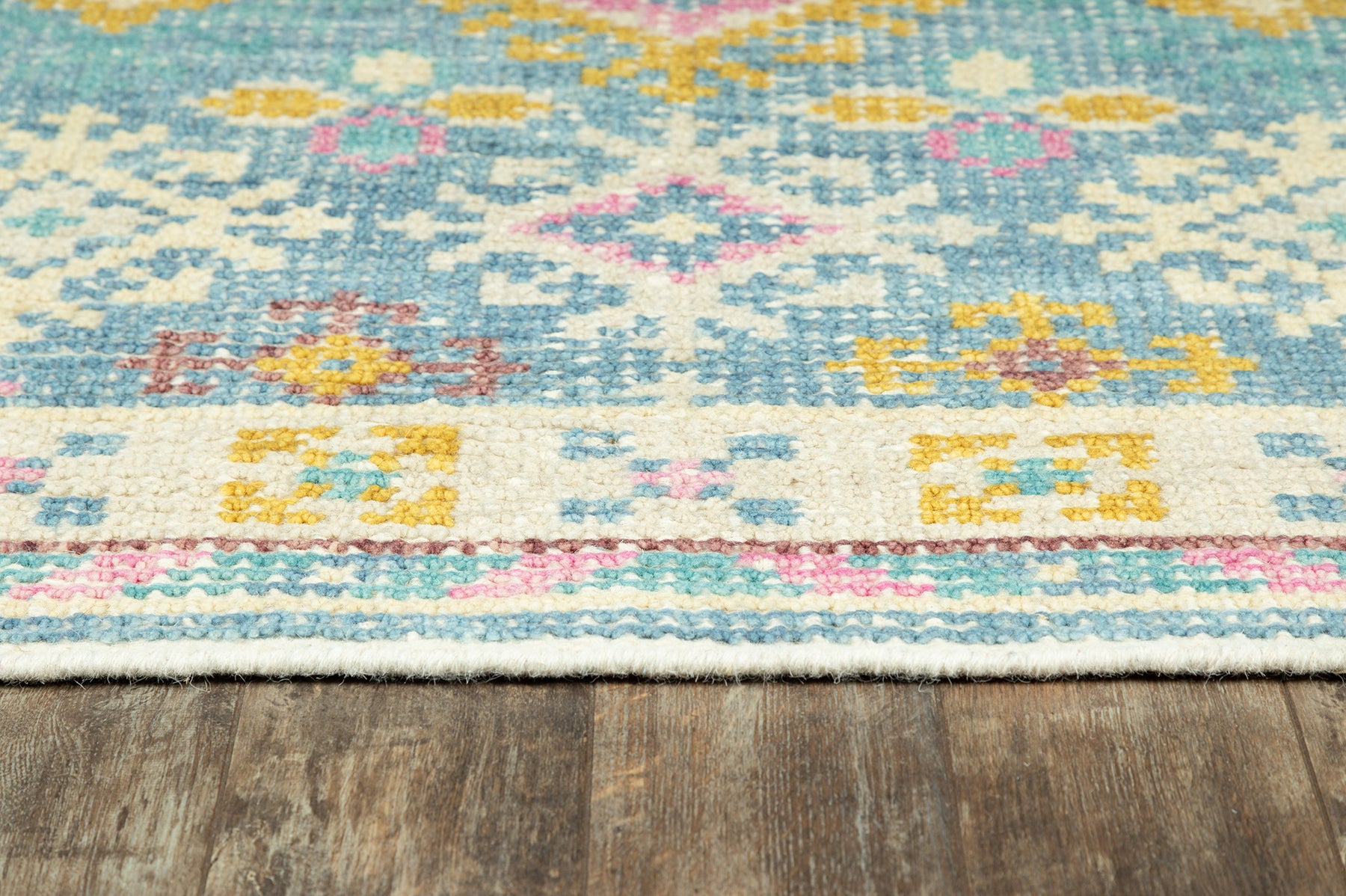 Turquoise kilim-style flat-weave rug with pink, yellow and cream geometric motifs and fringed edge on wood floor.