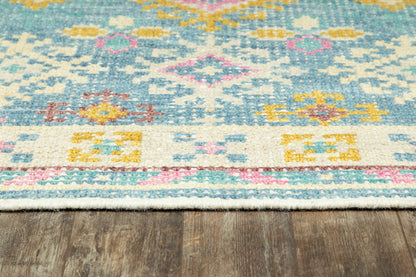 Turquoise kilim-style flat-weave rug with pink, yellow and cream geometric motifs and fringed edge on wood floor.