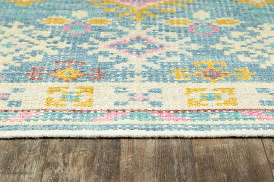 Turquoise kilim-style flat-weave rug with pink, yellow and cream geometric motifs and fringed edge on wood floor.