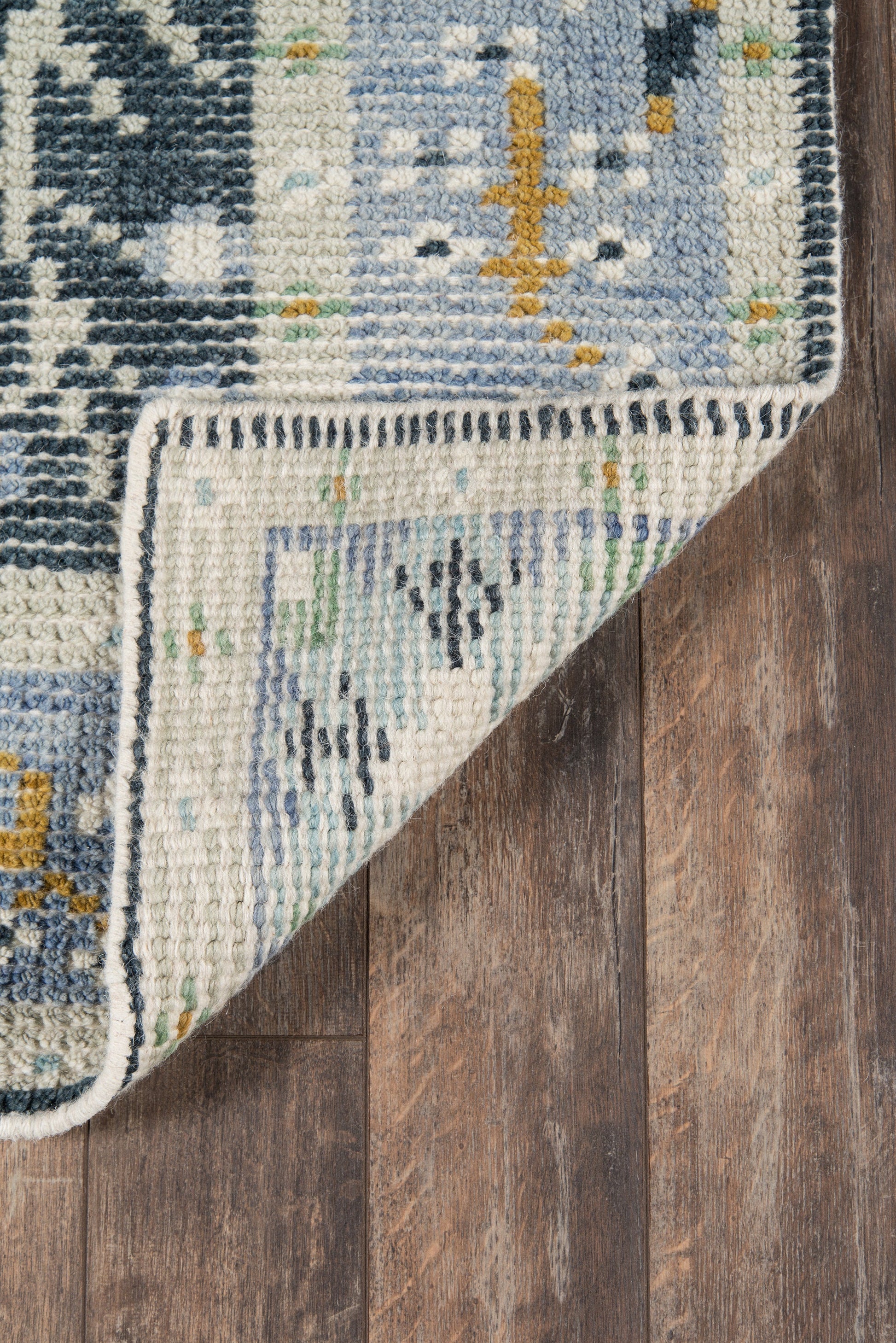Blue, ivory and teal geometric woven rug with mustard accents, folded to show a cream fringe border on wood floor.