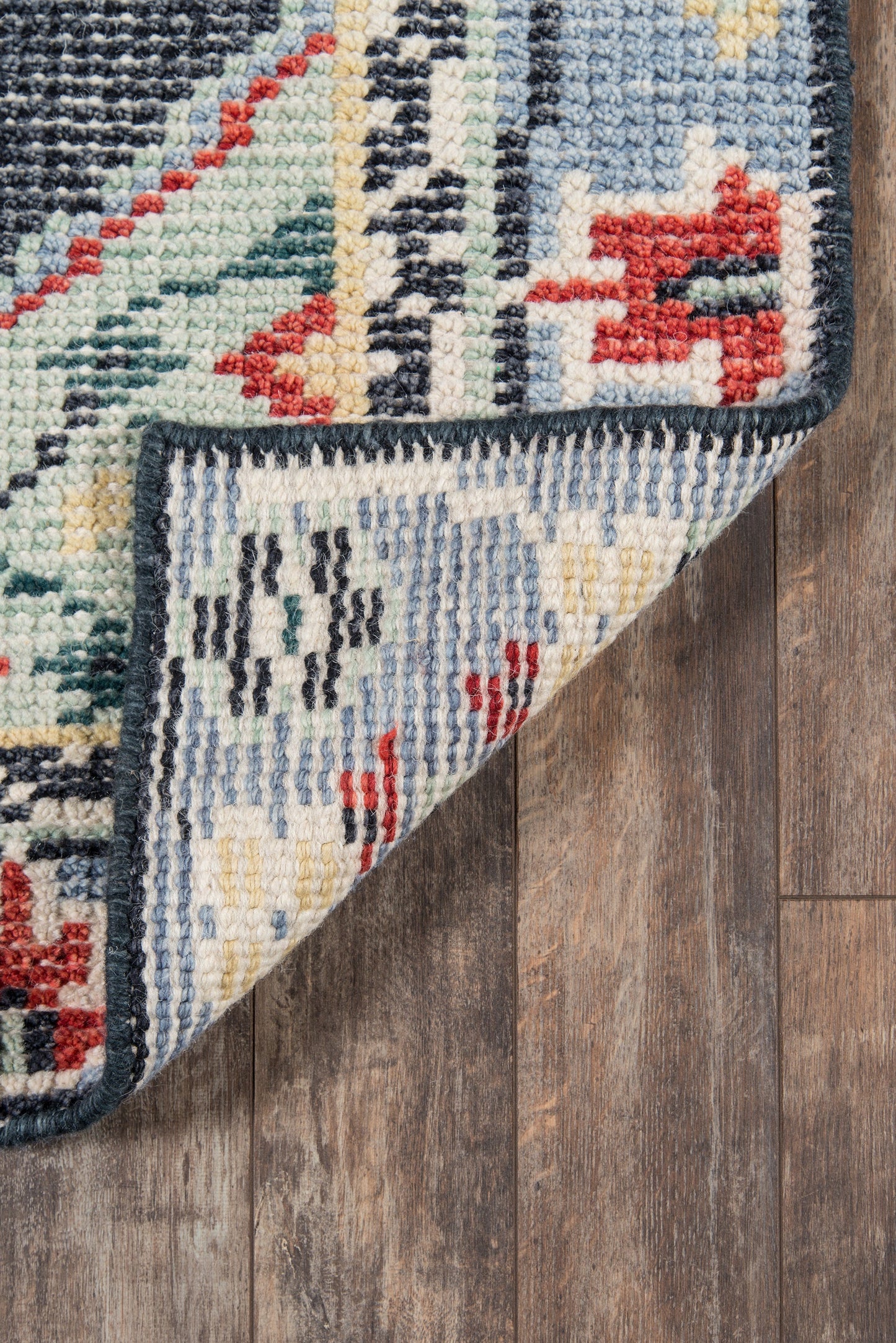 Woven kilim-style rug with geometric motifs in blues, cream, red & yellow; navy border, folded corner on wood floor.