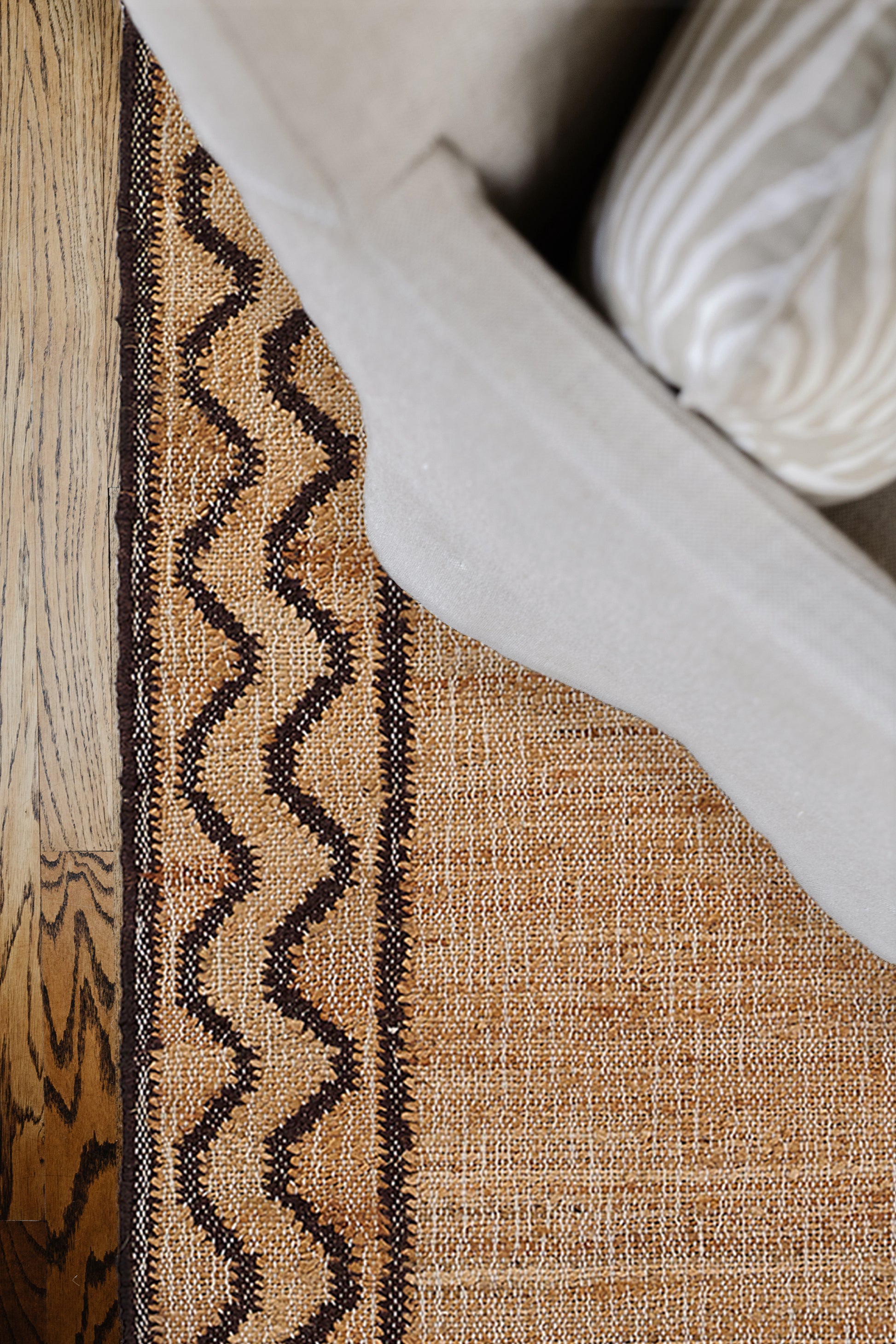 Beige and tan woven rug with a dark brown zigzag border, on a wood floor beside a gray sofa