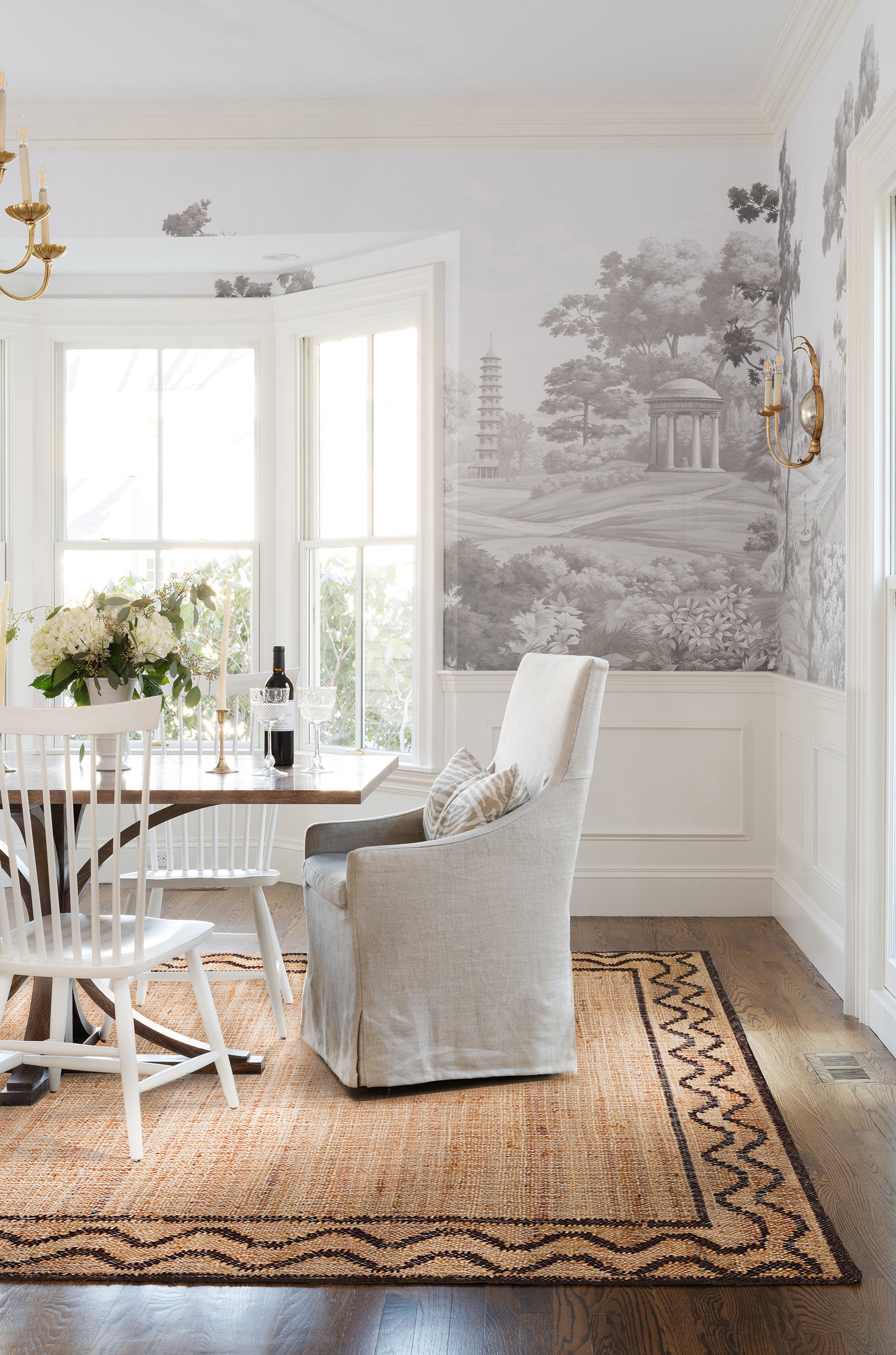 Bright dining nook with beige linen armchair, wooden table, white chairs, wine glasses and flowers on a rug, grayscale mural.