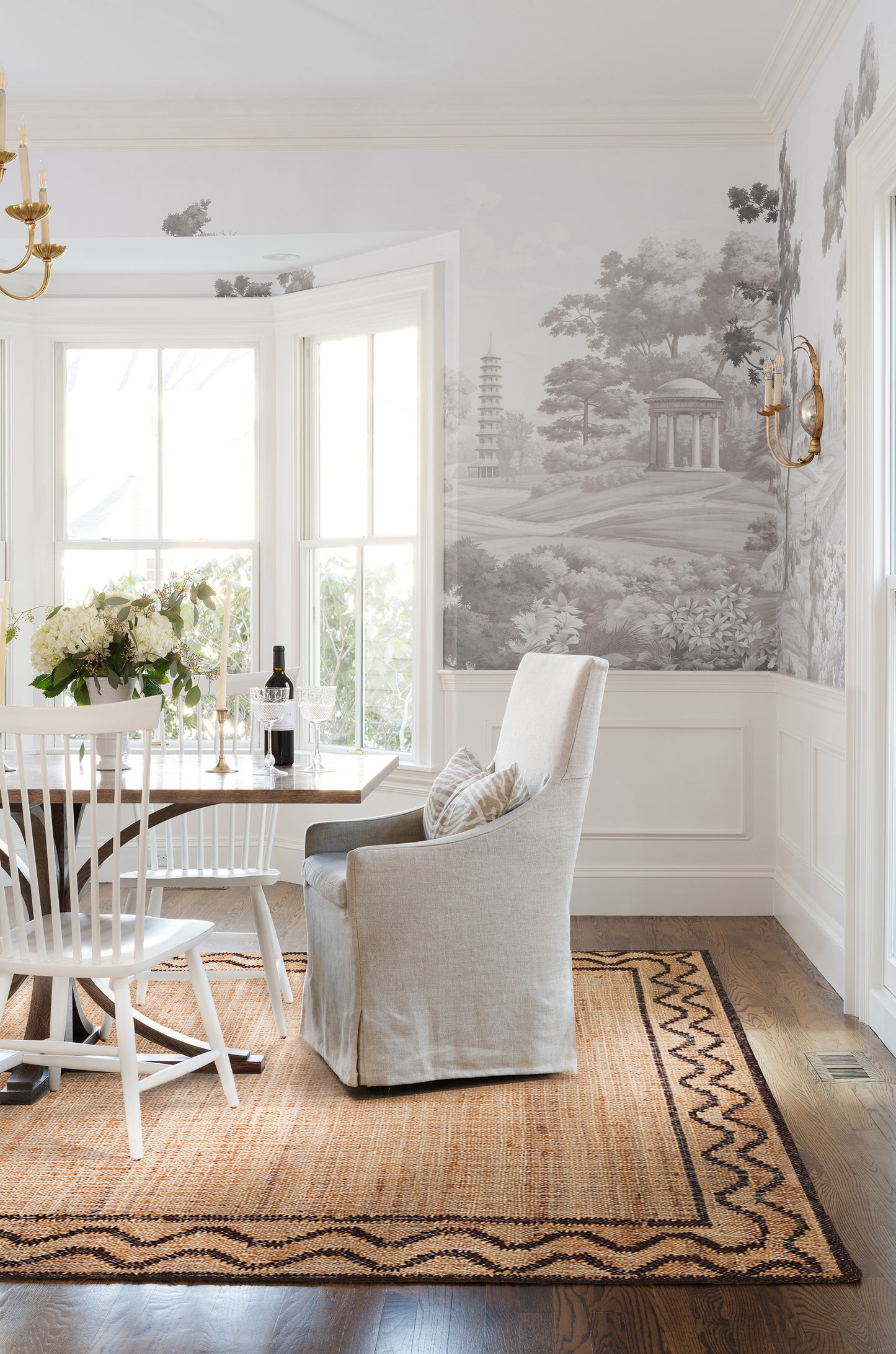 Bright dining nook with bay window, beige upholstered chair, white Windsor chairs, wooden table, jute rug, landscape mural.