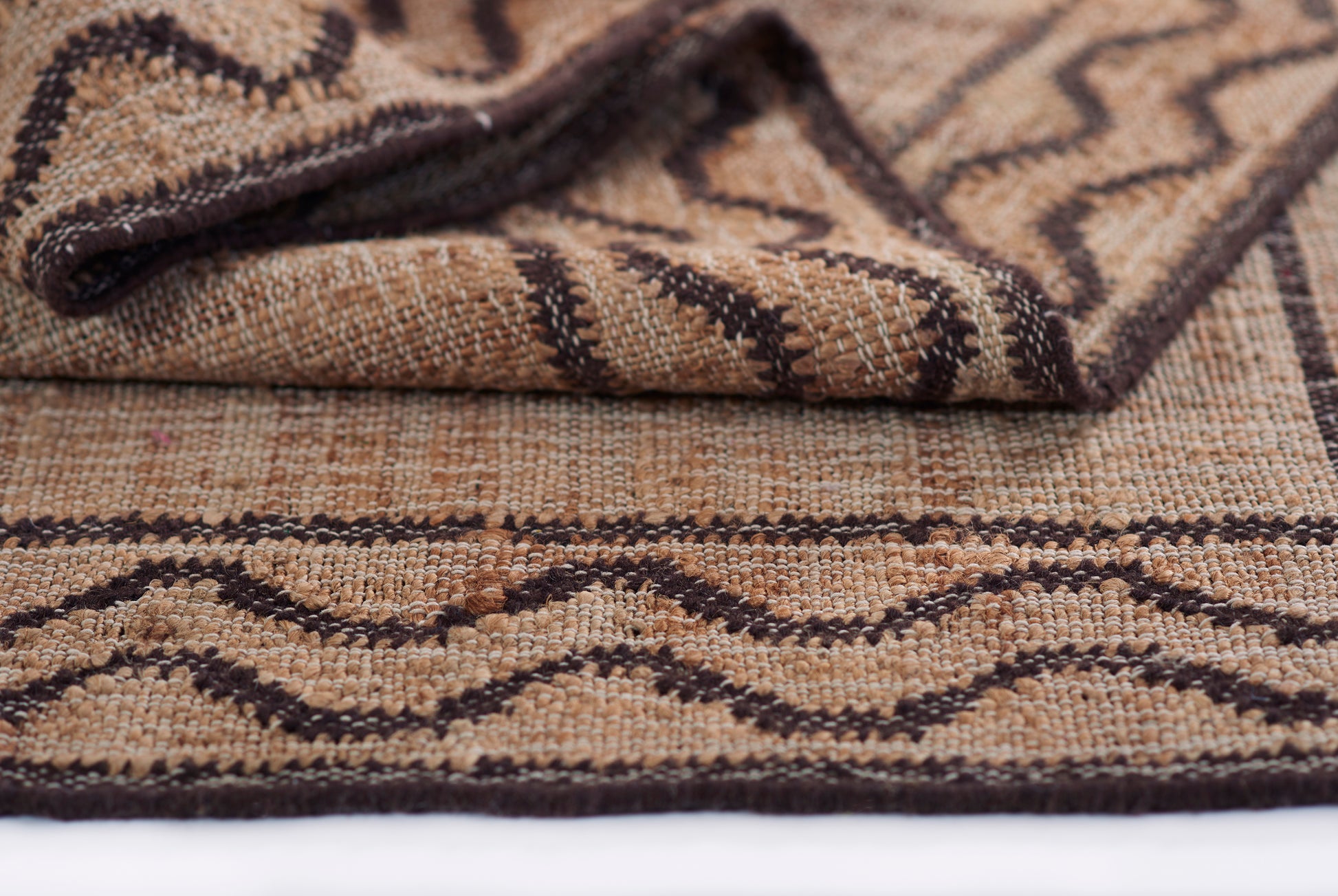 Natural beige-brown woven rug with dark brown chevron geometric pattern and folded corner.