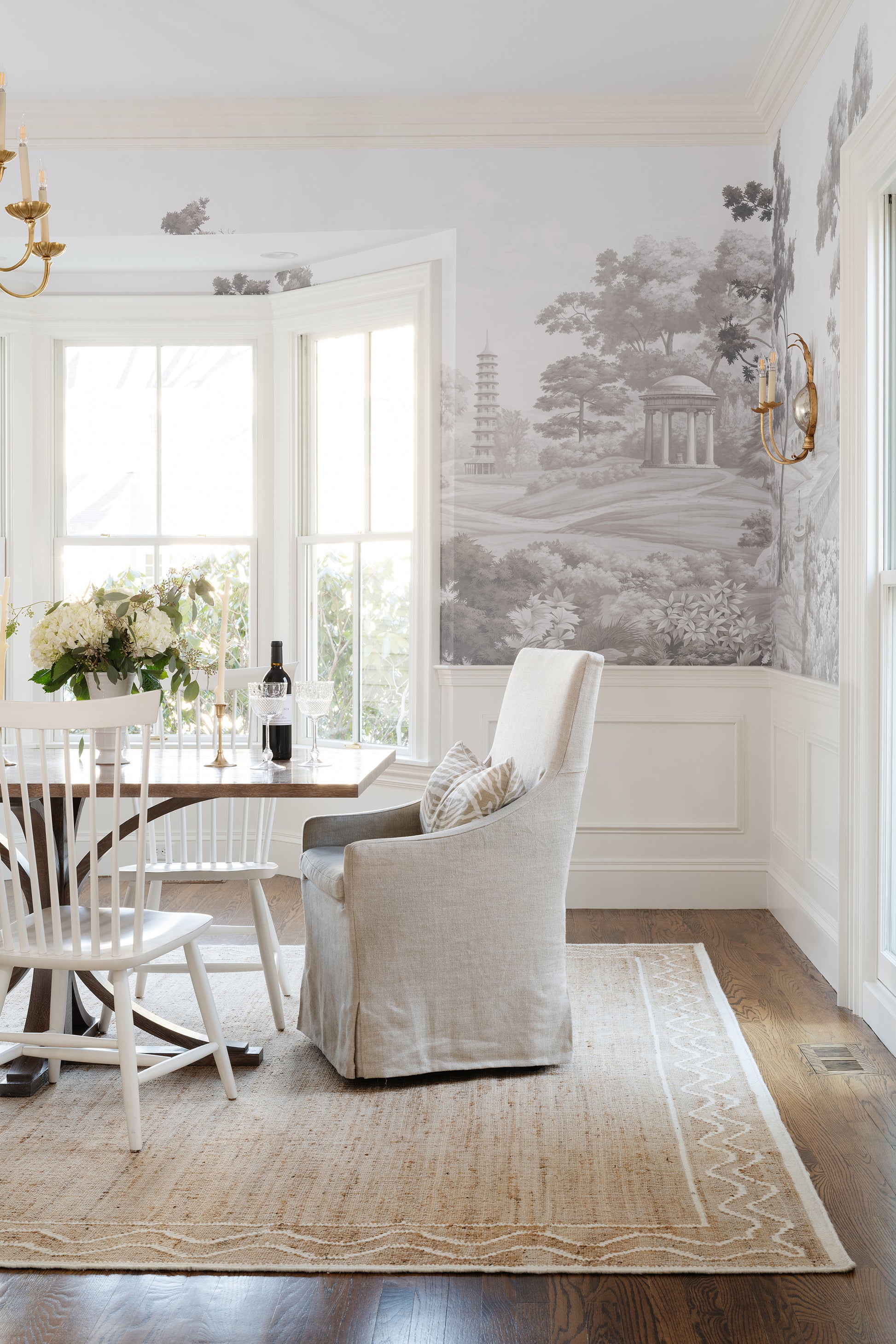 Bright dining room with a rug under a wood table and white chairs, floral centerpiece, beige armchair, grayscale landscape wallpaper, bay window.