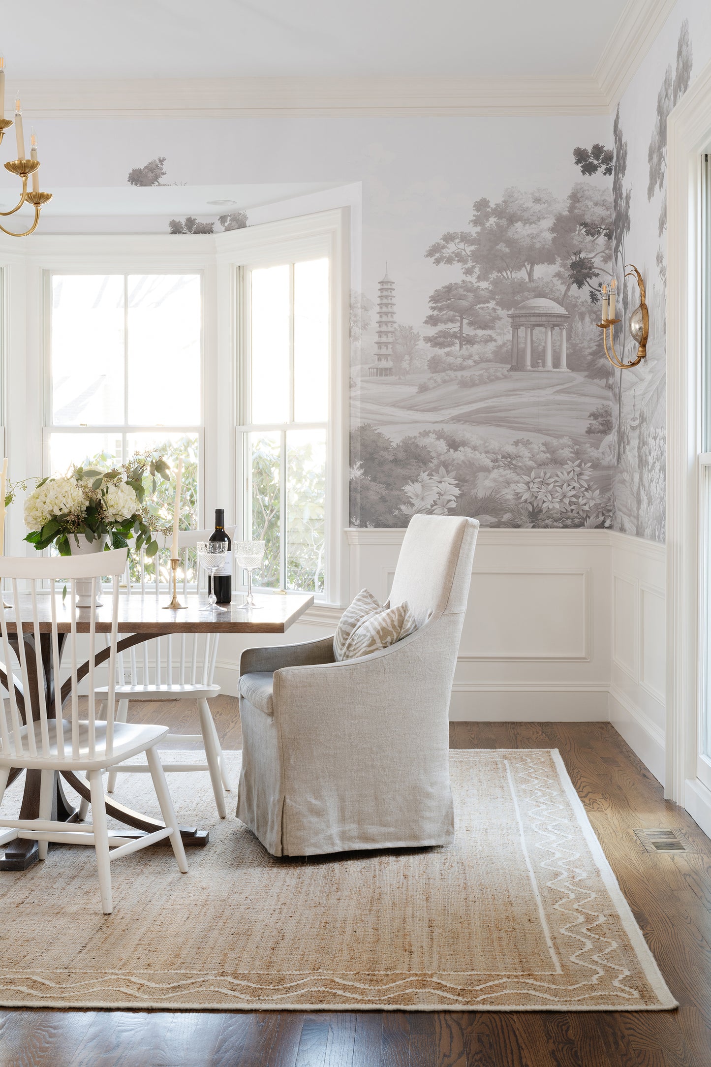 Neutral dining room with beige armchair, wooden table, white chairs, black-and-white mural, jute rug, wine centerpiece