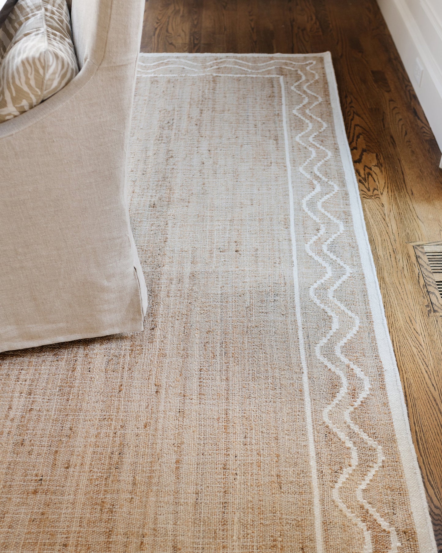 Natural beige jute rug with a white wavy border along one edge, beside a beige sofa on dark hardwood floor.