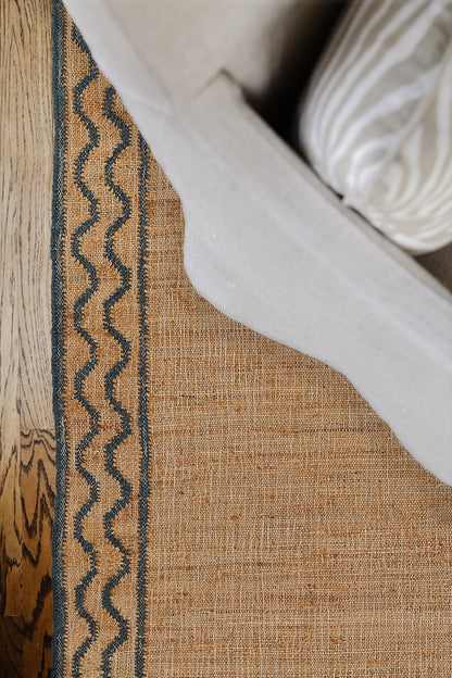 Warm orange-brown woven rug with a dark blue zigzag border beside a light gray sofa on a wooden floor.