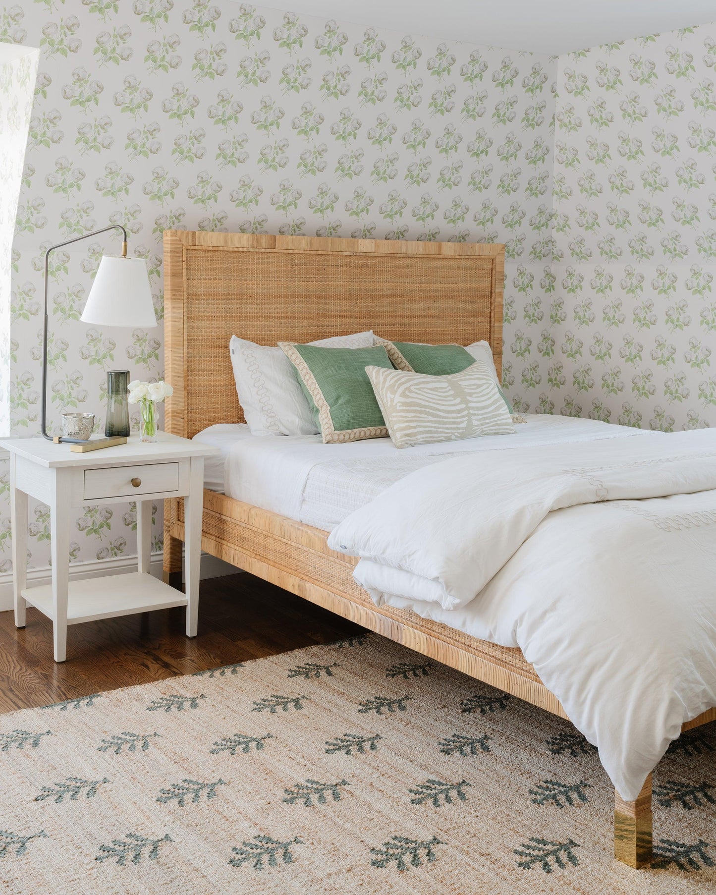 Natural rattan bed frame with white duvet, green and beige cushions; white nightstand, leafy wallpaper, and patterned rug.