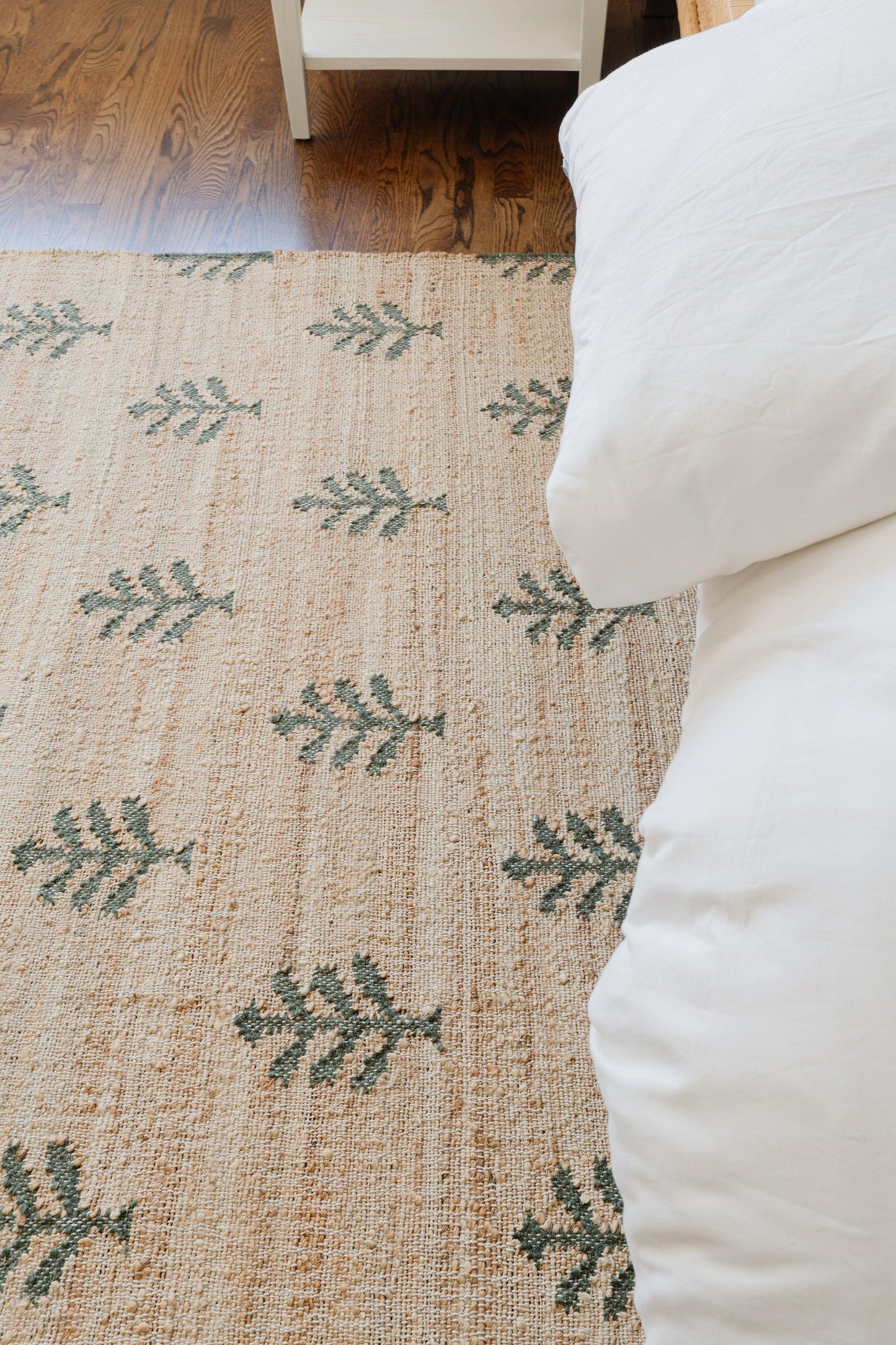 Natural jute rug with green fern leaf motifs on a hardwood floor beside white bedding; rectangular boho bedroom rug.