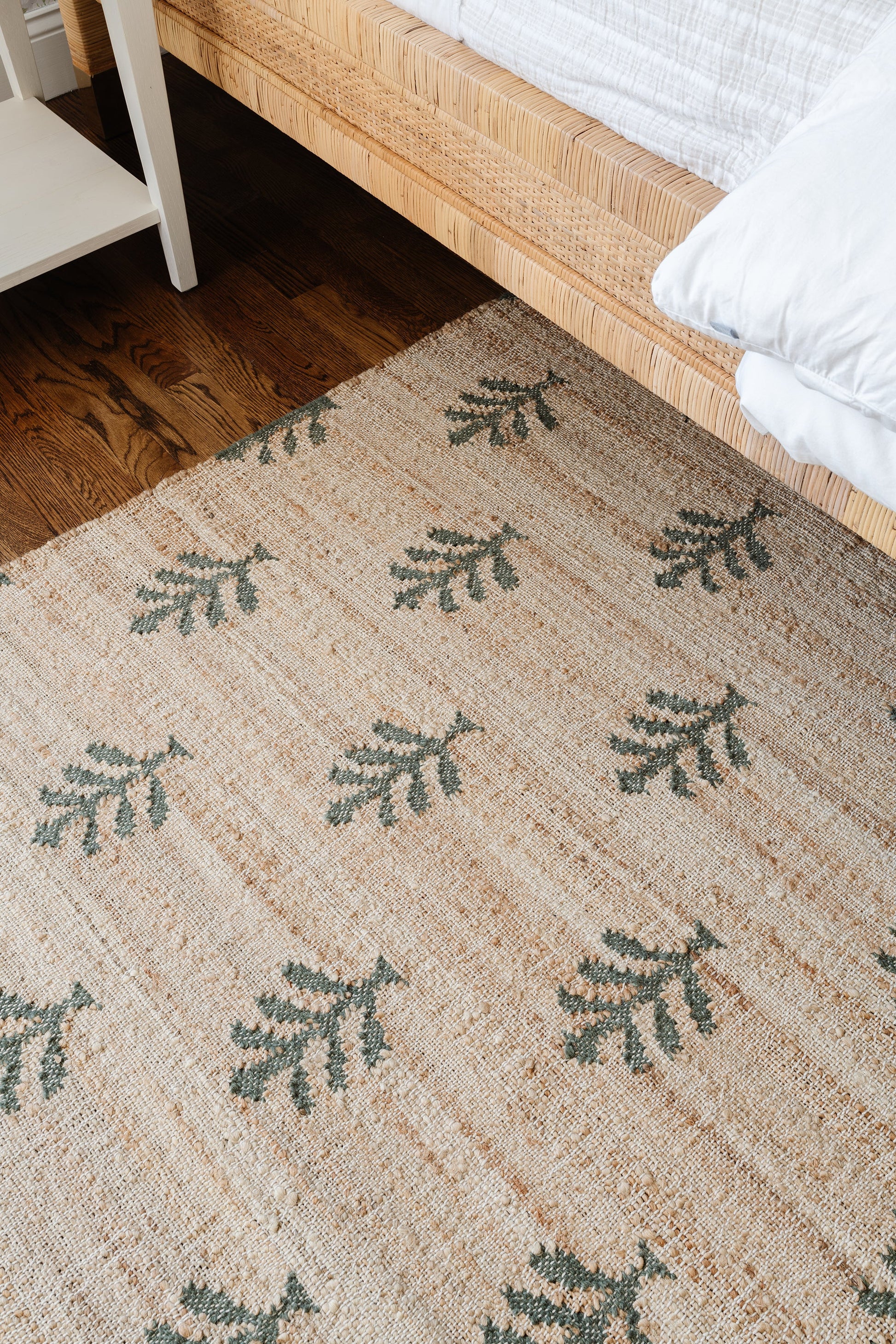 Natural jute rug with green fern motifs beside a woven rattan bed frame on dark wood flooring.