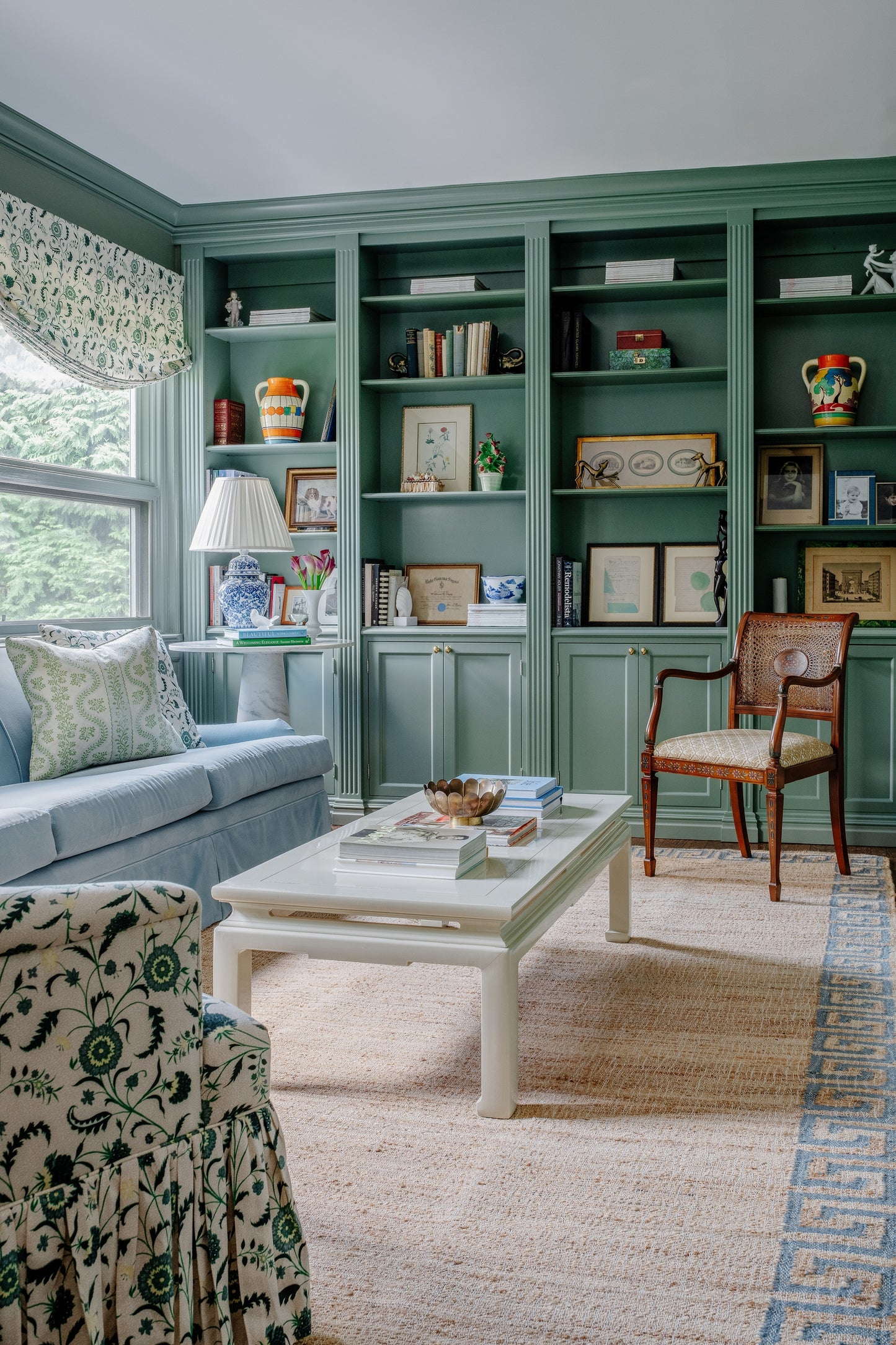 Teal built-in shelves with display cabinets flank a cozy living room; blue sofa, white coffee table, wooden chair, beige rug.
