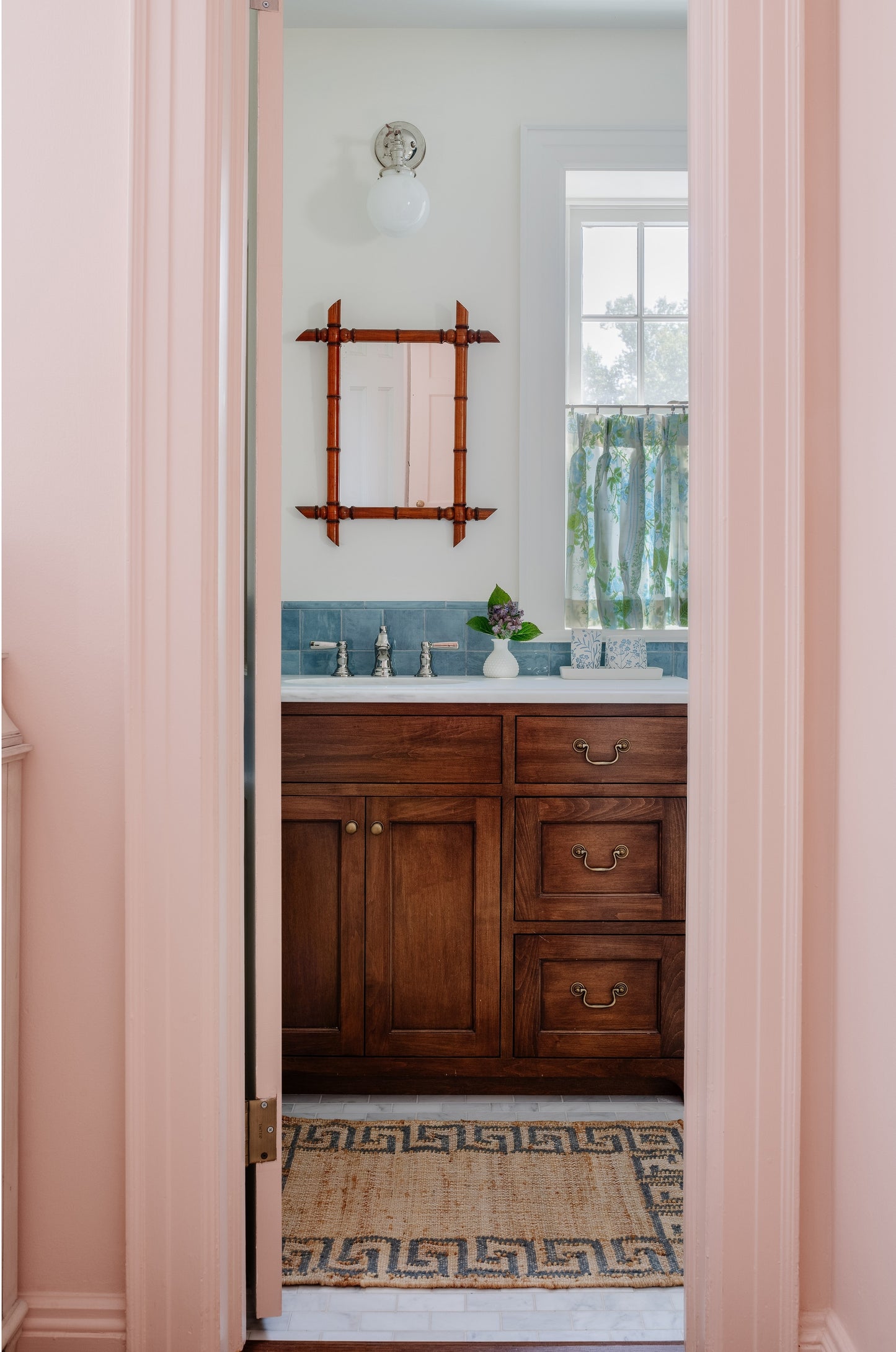 Pink doorway opens to a bath with dark wood vanity, blue tile, bamboo-framed mirror, floral curtain, Greek-key rug.