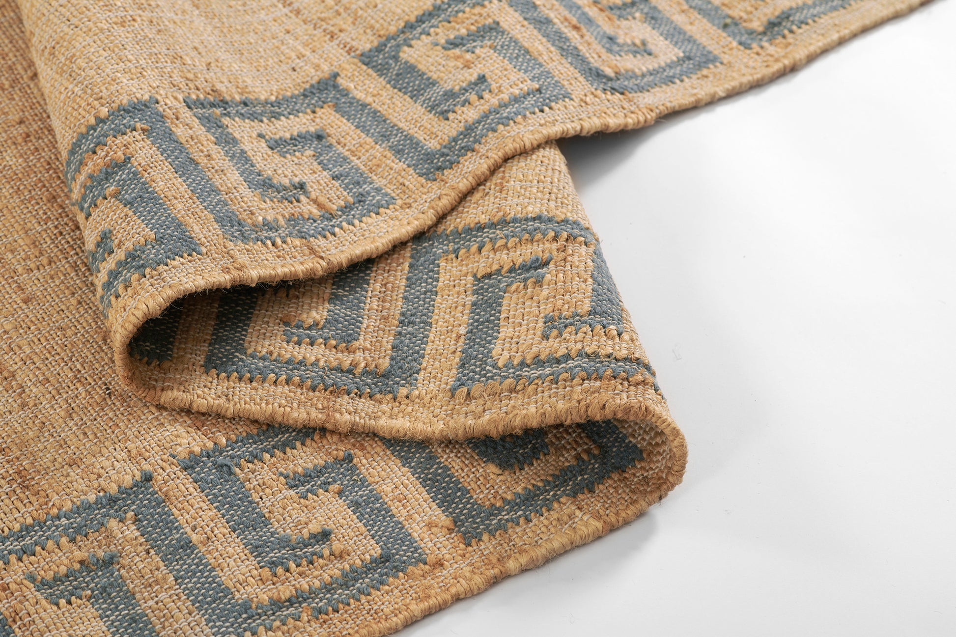 Beige woven rug with blue Greek-key border, folded corner shows textured natural fibers.