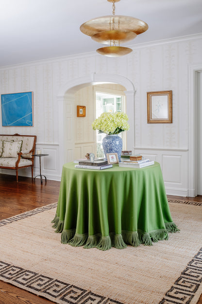Round table draped in emerald green velvet with fringe, blue-and-white vase of hydrangeas, stacked books in a traditional living room.
