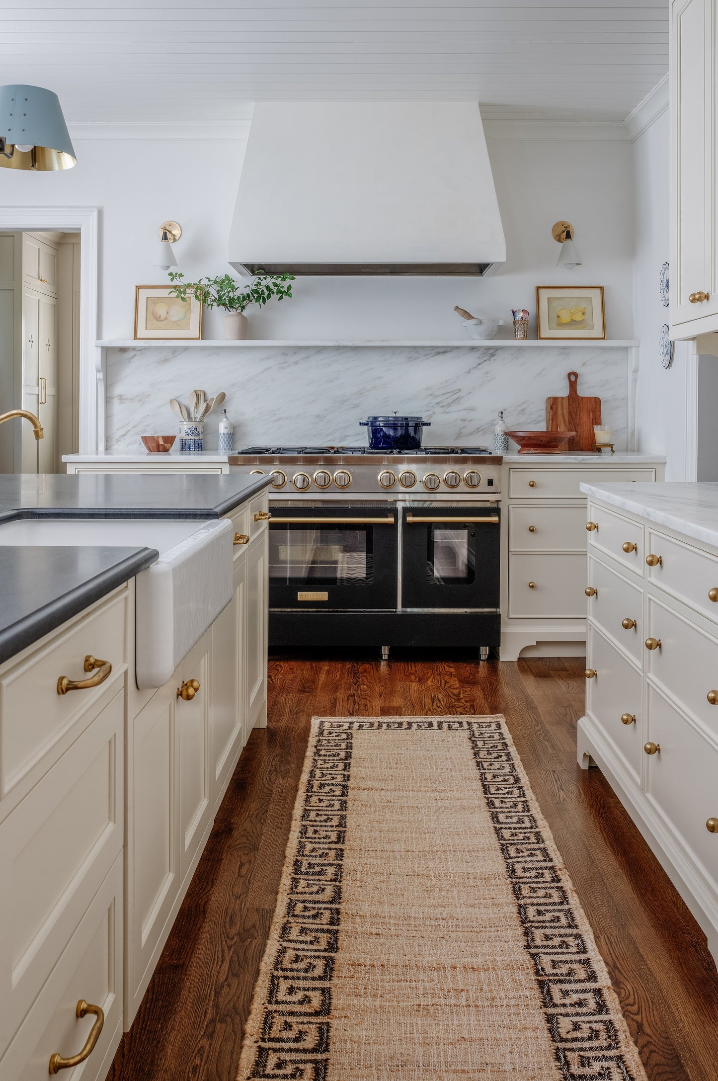 White kitchen with marble backsplash, brass hardware, black 6-burner range, white hood, dark wood floor and a beige runner.