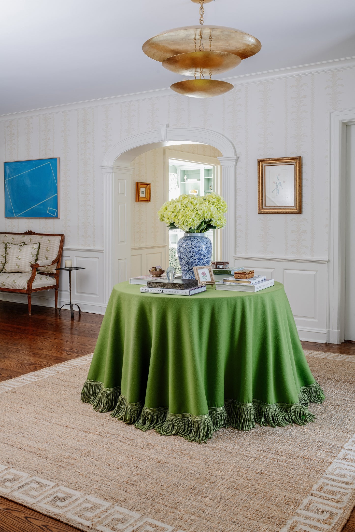 Living room with a Greek key patterned rug runner on the floor; round table with green velvet fringe, blue-and-white vase of white hydrangeas, arched doorway and brass chandelier.