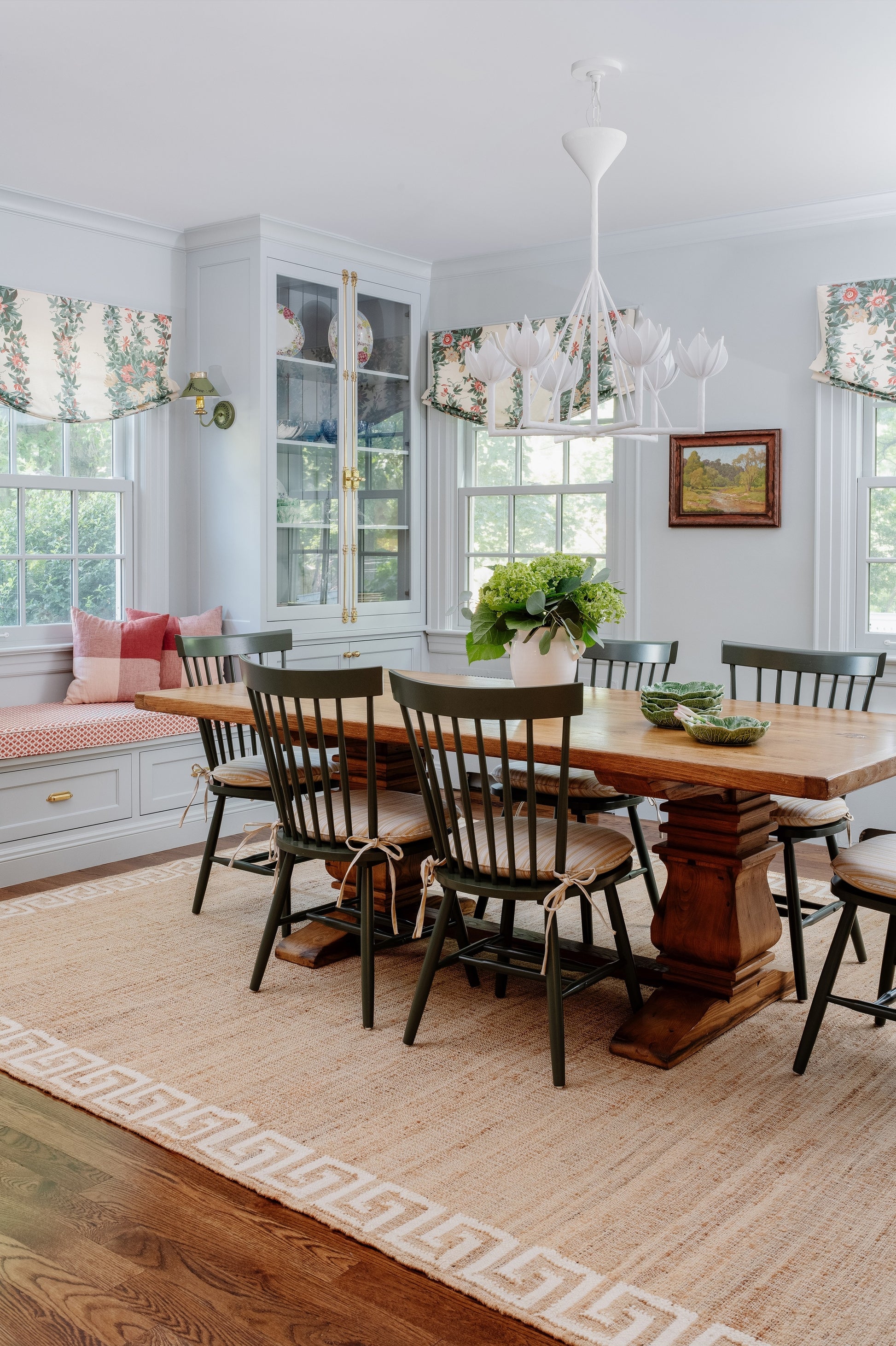 Bright blue dining room with a solid wood farmhouse table, black Windsor chairs, woven rug, and glass-front cabinets.