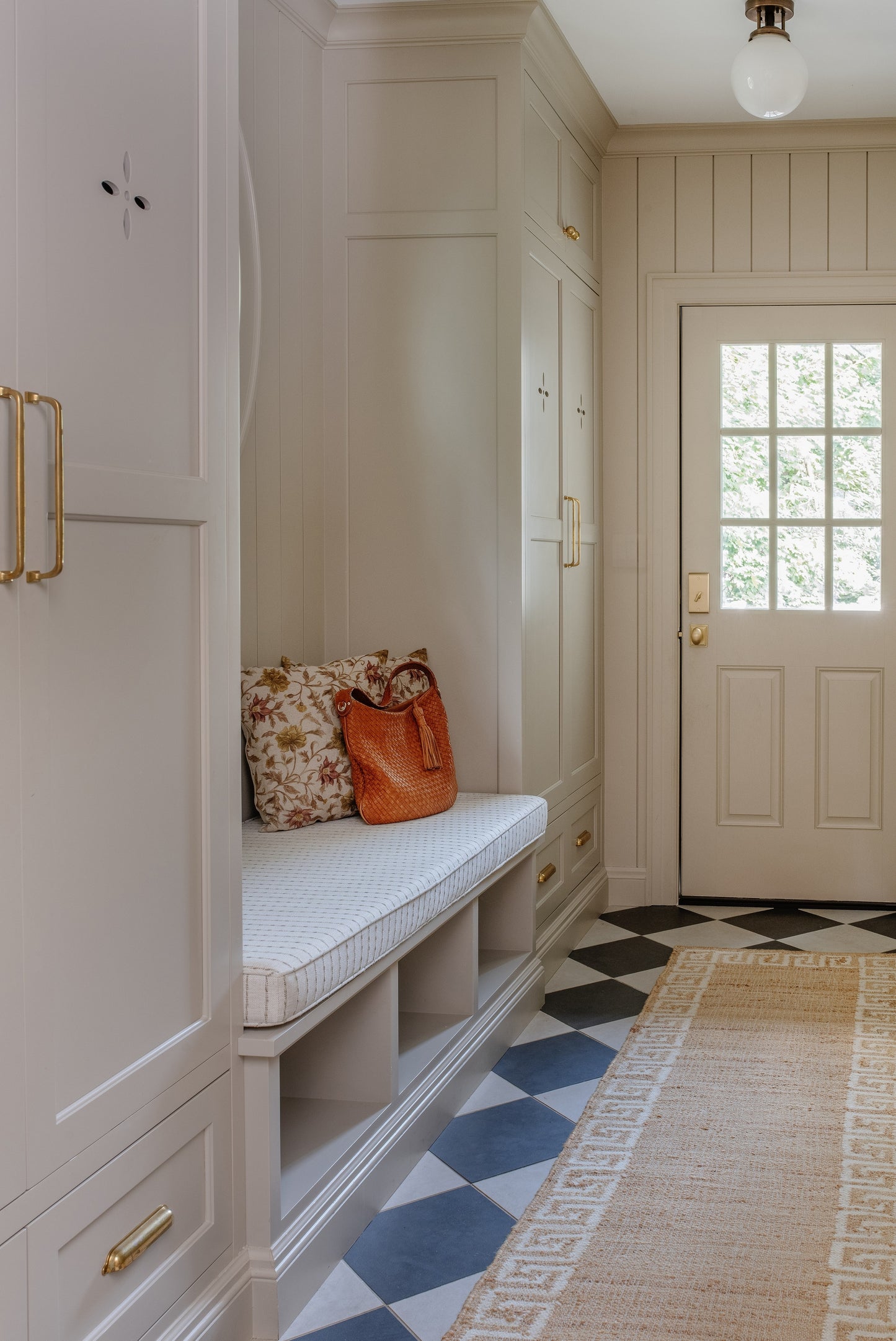 White mudroom with bench, gold hardware, floral cushion, orange bag, blue-and-white checkered floor, woven rug, front door