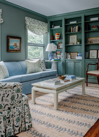Teal built-in shelves, light blue sofa, floral armchair, white coffee table, striped rug in a coastal-traditional living room.
