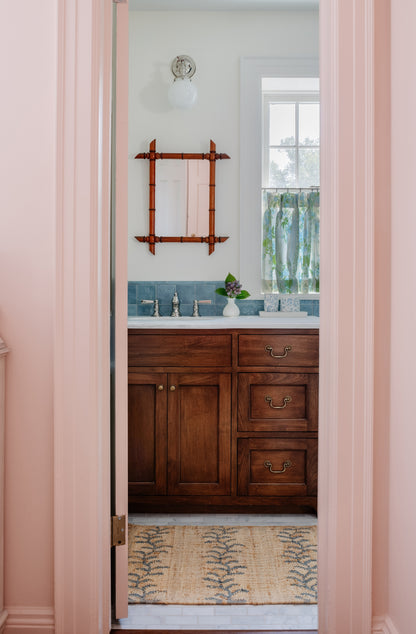 Rustic dark-wood bathroom vanity with light countertop, brass pulls, blue backsplash, framed mirror, pink doorway frame.