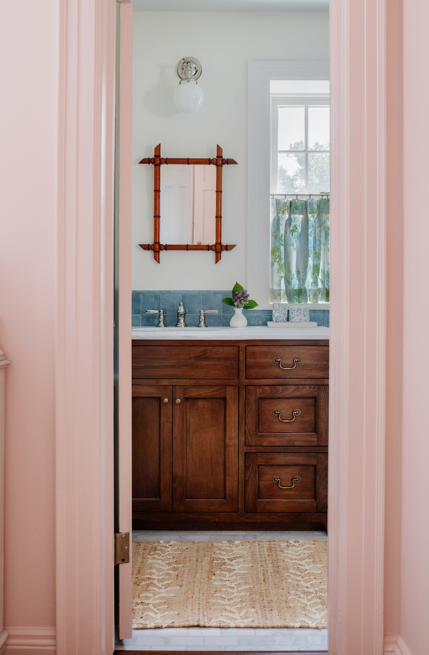 Pink doorway into a bathroom with dark wood vanity, blue tile backsplash, bamboo-framed mirror, globe light, vase of flowers.