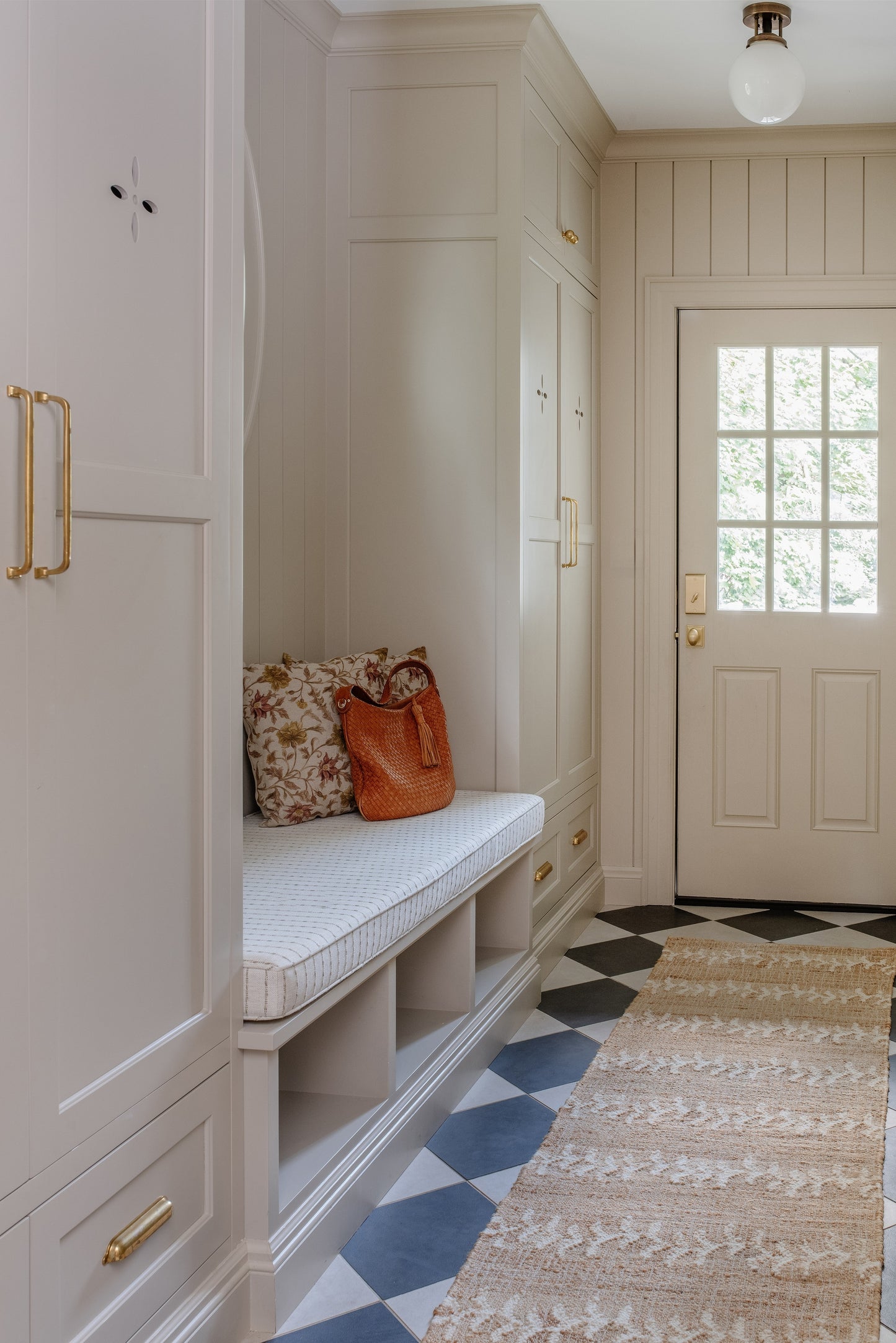 White built-in entry with bench and cubbies, brass handles, black-and-white floor tiles, beige rug, floral pillow and orange bag.
