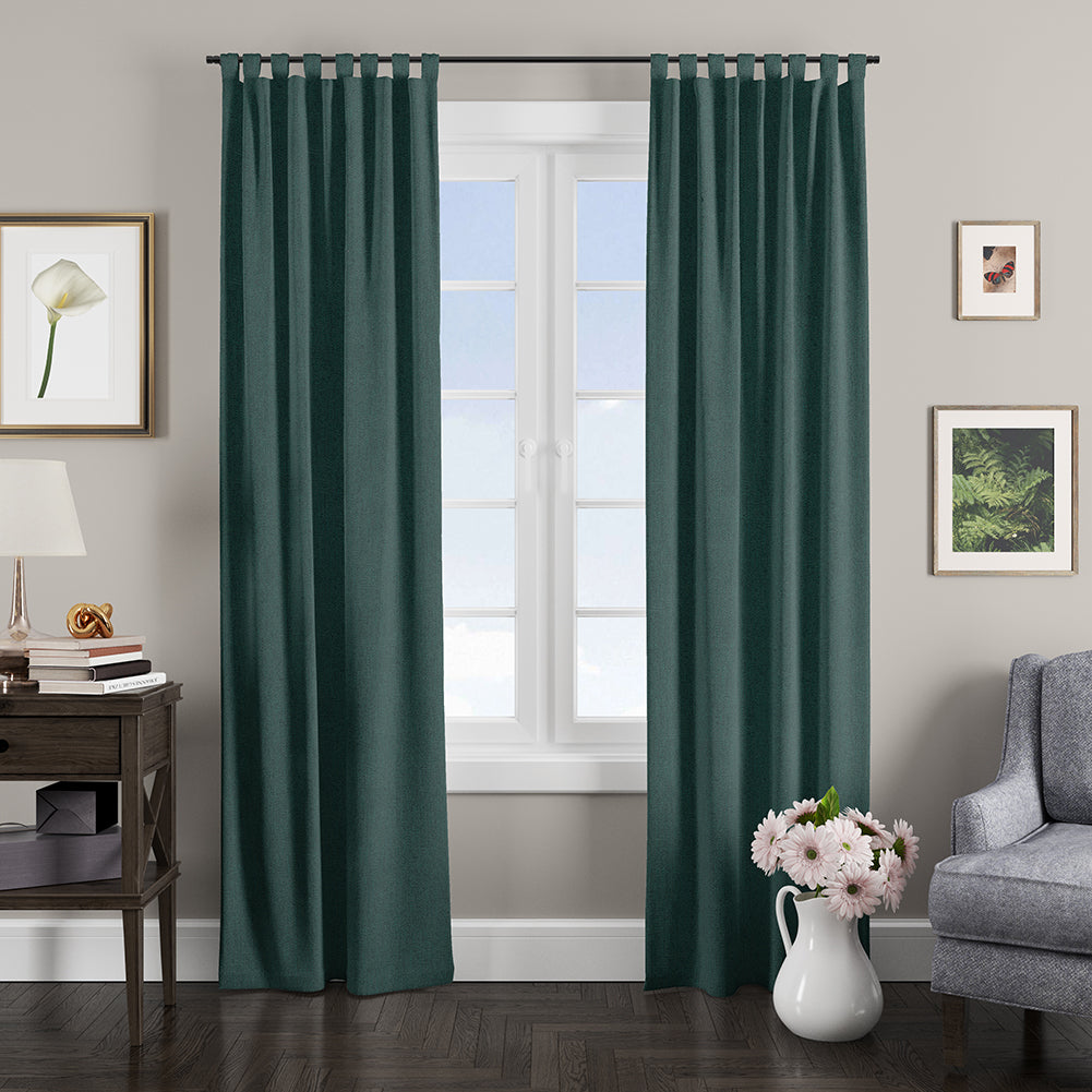 Emerald teal curtains frame a bright window in a cozy living room with a side table, lamp, art, gray sofa, and pink flowers.