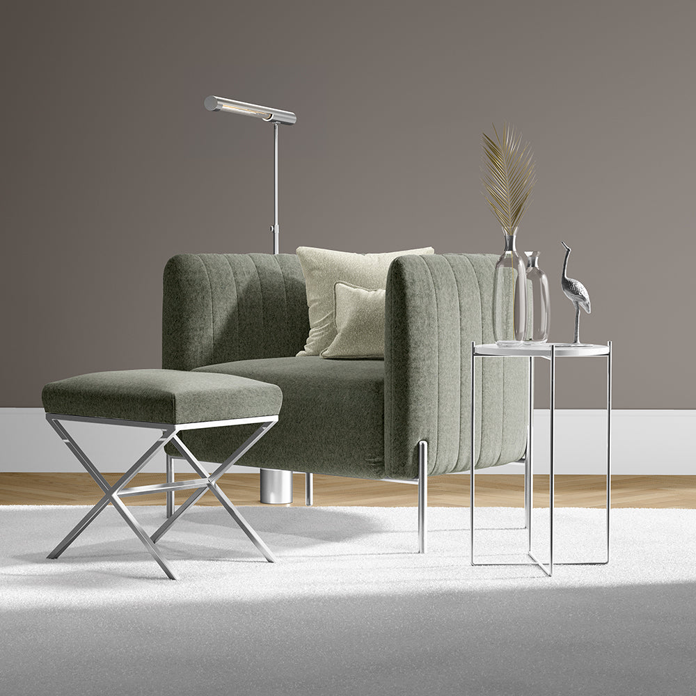 Olive green velvet sofa with matching ottoman, chrome legs; glass side tables with vases and dried plant, modern floor lamp.